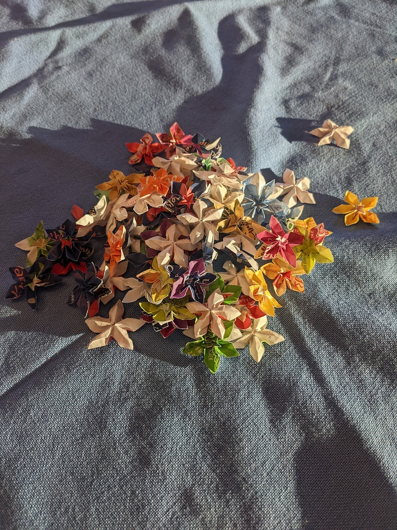 May include: A collection of colorful origami flowers made from paper. The flowers are arranged in a pile on a blue fabric surface.