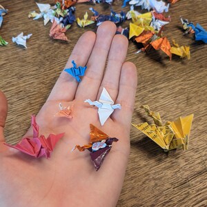 Może przedstawiać: Ręka trzymająca kilka origami z papieru w kształcie żurawi. Żurawie są wykonane z papieru w r&oacute;żnych kolorach, w tym czerwonym, niebieskim, białym, ż&oacute;łtym i pomarańczowym. Żurawie są złożone w tradycyjnym japońskim stylu.