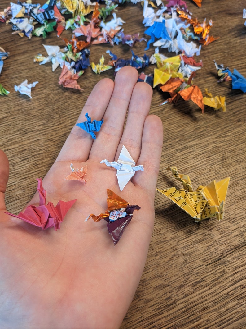 Może przedstawiać: Ręka trzymająca kilka origami z papieru w kształcie żurawi. Żurawie są wykonane z papieru w r&oacute;żnych kolorach, w tym czerwonym, niebieskim, białym, ż&oacute;łtym i pomarańczowym. Żurawie są złożone w tradycyjnym japońskim stylu.