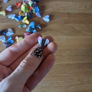 May include: A hand holding a small, folded paper heart with a black and white striped pattern. The heart is made of paper and has a white background with black stripes. The hand is wearing nail polish with a colorful polka dot pattern.