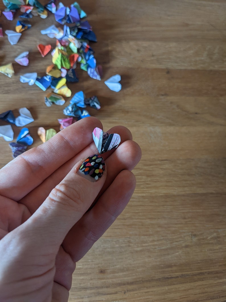 May include: A hand holding a small, folded paper heart with a black and white striped pattern. The heart is made of paper and has a white background with black stripes. The hand is wearing nail polish with a colorful polka dot pattern.