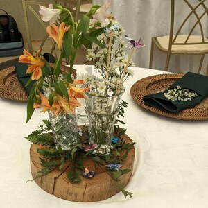 May include: A wooden slice with green foliage and two glass vases filled with white and orange flowers. The vases are decorated with small butterflies. The wooden slice is on a white tablecloth with two wicker plates with green napkins and white flowers.