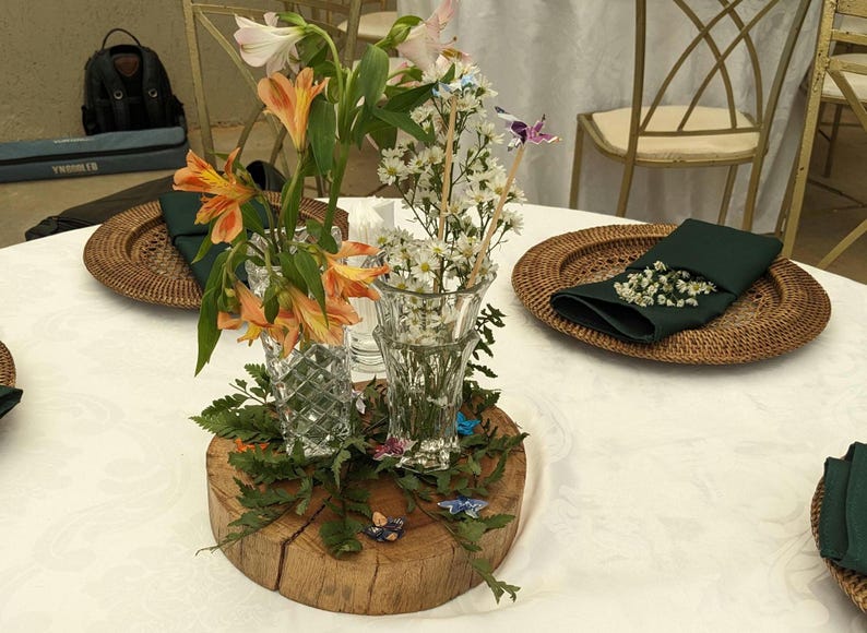 May include: A wooden slice with green foliage and two glass vases filled with white and orange flowers. The vases are decorated with small butterflies. The wooden slice is on a white tablecloth with two wicker plates with green napkins and white flowers.
