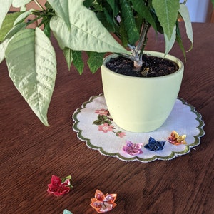 May include: A potted plant with green leaves sits on a table with a white doily.  There are six origami flowers on the table, in colors of red, pink, blue, green, orange, and yellow.