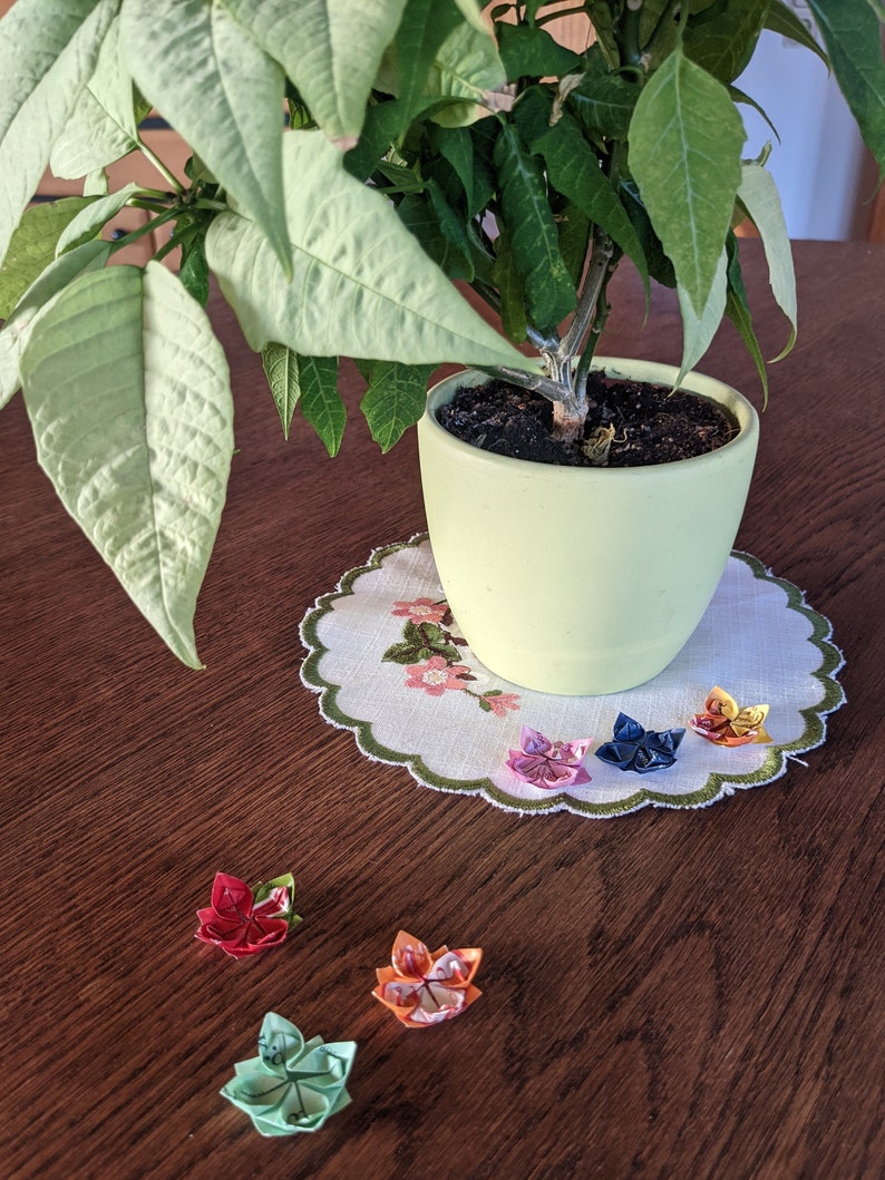 May include: A potted plant with green leaves sits on a table with a white doily.  There are six origami flowers on the table, in colors of red, pink, blue, green, orange, and yellow.