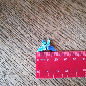 May include: A small blue origami crane made from paper, measured against a red ruler. The crane is approximately 2 cm wide.