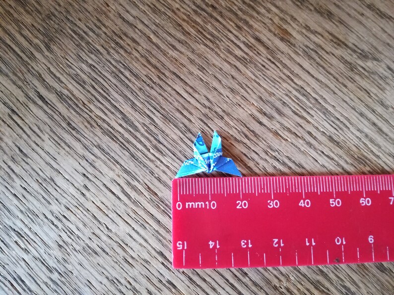May include: A small blue origami crane made from paper, measured against a red ruler. The crane is approximately 2 cm wide.