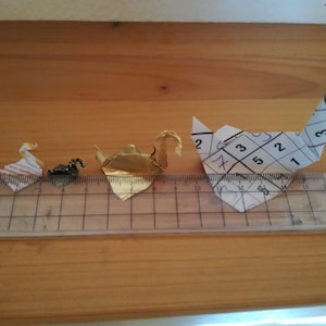 May include: Three origami paper cranes of different sizes. The largest crane is white with black markings and is made from a piece of paper with numbers written on it. The middle crane is gold and the smallest crane is white and red.