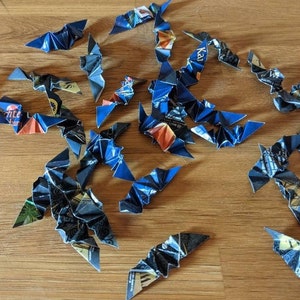 May include: A collection of origami bats made from recycled blue and black paper. The bats are scattered across a wooden surface, showcasing their folded wing details and various patterns from the original paper.