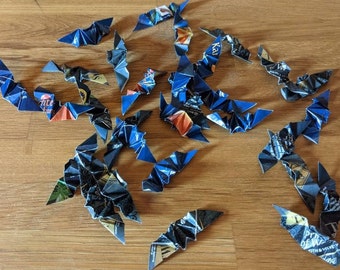Puede incluir: Un conjunto de murciélagos de origami hechos con papel reciclado azul y negro. Los murciélagos están dispersos sobre una superficie de madera, mostrando los detalles de sus alas plegadas y los patrones del papel original.
