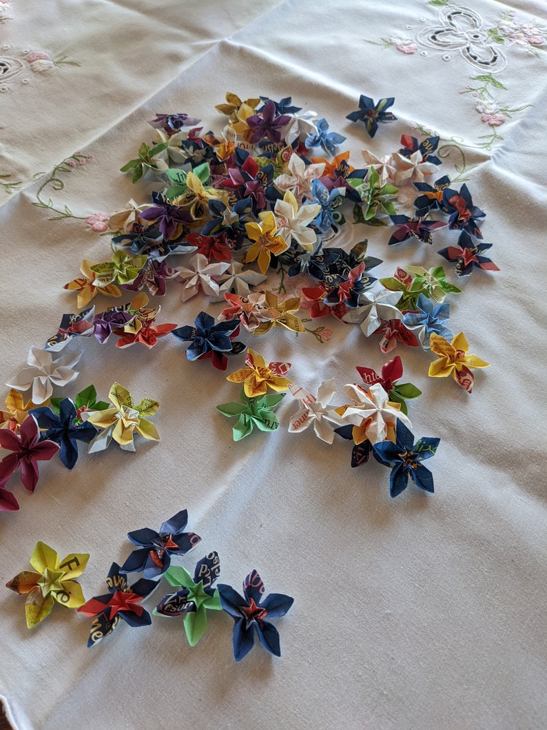 May include: A collection of colorful origami flowers made from paper. The flowers are arranged on a white tablecloth with a floral pattern.