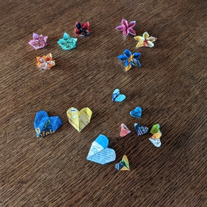 May include: A collection of origami flowers and hearts made from colorful paper. The flowers are in various colors, including pink, blue, yellow, and red. The hearts are mostly blue and yellow.