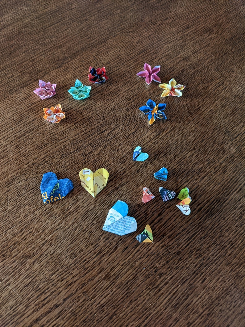May include: A collection of origami flowers and hearts made from colorful paper. The flowers are in various colors, including pink, blue, yellow, and red. The hearts are mostly blue and yellow.