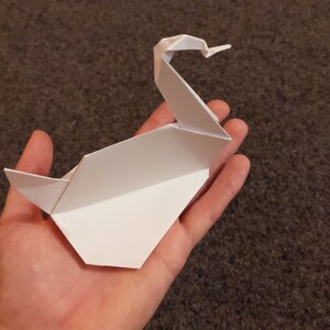 May include: A white origami swan with a long neck and a curved beak. The swan is folded from a single sheet of paper and is sitting in the palm of a hand.