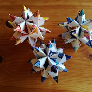 Puede incluir: Tres estrellas de origami coloridas hechas de papel reciclado, con una variedad de patrones y colores. Las estrellas est&aacute;n dispuestas sobre una superficie de madera, mostrando sus formas geom&eacute;tricas y pliegues intrincados. Un proyecto de manualidades creativo y ecol&oacute;gico.