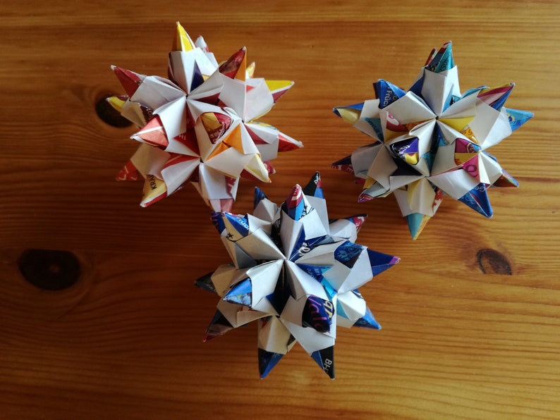 Puede incluir: Tres estrellas de origami coloridas hechas de papel reciclado, con una variedad de patrones y colores. Las estrellas est&aacute;n dispuestas sobre una superficie de madera, mostrando sus formas geom&eacute;tricas y pliegues intrincados. Un proyecto de manualidades creativo y ecol&oacute;gico.
