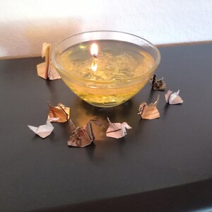 May include: A clear glass bowl filled with a yellow liquid and a lit candle inside. The bowl is surrounded by several small origami paper cranes in various colors.