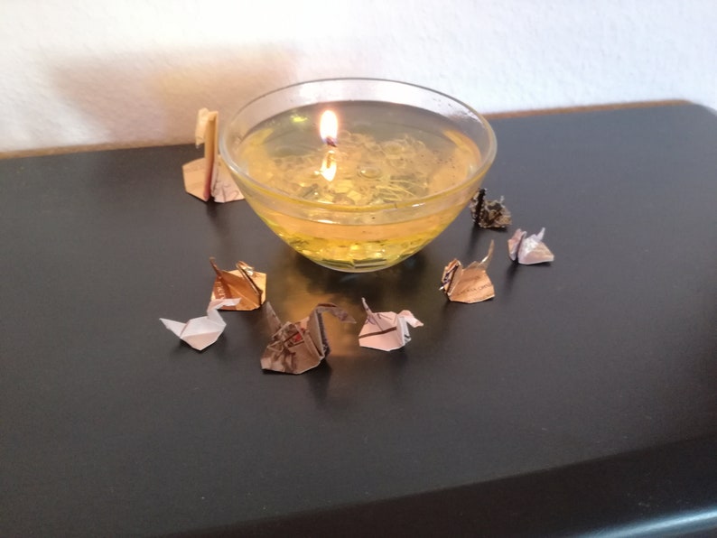 May include: A clear glass bowl filled with a yellow liquid and a lit candle inside. The bowl is surrounded by several small origami paper cranes in various colors.