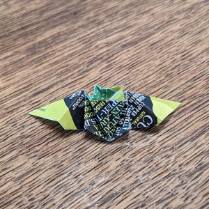 Puede incluir: Una peque&ntilde;a mariposa de origami hecha de un envoltorio de bolsita de t&eacute;, con texto verde, amarillo y negro. La mariposa est&aacute; sobre una superficie de madera, mostrando la marca de t&eacute;.