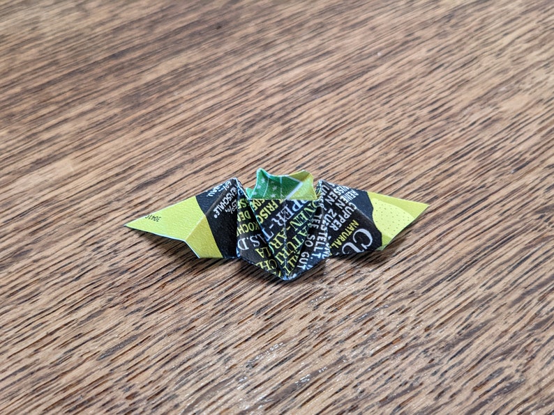Puede incluir: Una peque&ntilde;a mariposa de origami hecha de un envoltorio de bolsita de t&eacute;, con texto verde, amarillo y negro. La mariposa est&aacute; sobre una superficie de madera, mostrando la marca de t&eacute;.