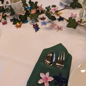 May include: A green napkin folded into a pocket with a silver spoon and fork inside. The napkin is decorated with origami flowers in pink, white, and blue.