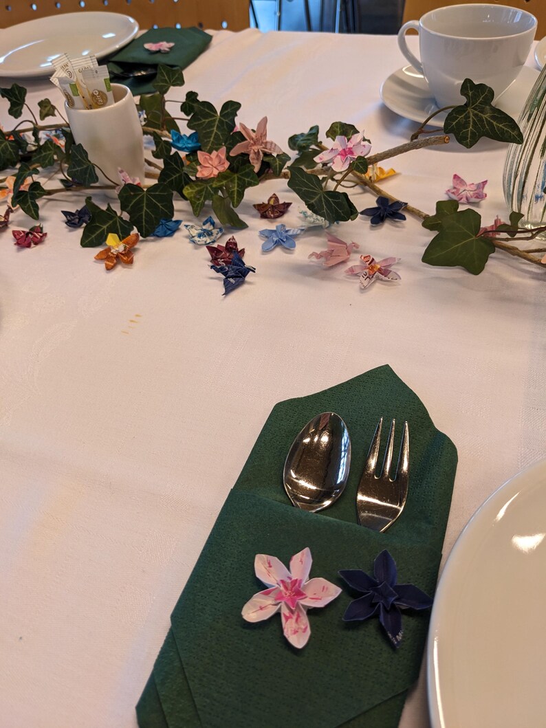 May include: A green napkin folded into a pocket with a silver spoon and fork inside. The napkin is decorated with origami flowers in pink, white, and blue.