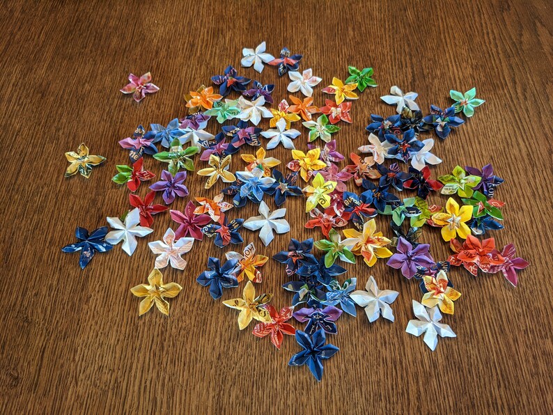 May include: A collection of colorful origami flowers made from paper. The flowers are in various shades of blue, green, yellow, orange, red, and white. The flowers are arranged in a loose cluster on a wooden surface.