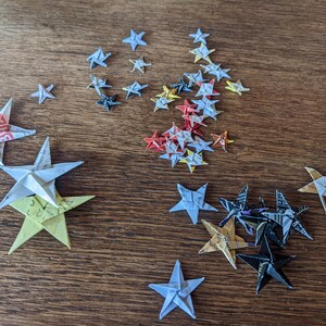 Puede incluir: Estrellas de origami de varios tama&ntilde;os y colores, incluyendo blanco, amarillo, rojo y negro. Las estrellas est&aacute;n dispuestas sobre una superficie de madera, mostrando los intrincados pliegues y patrones del papel.