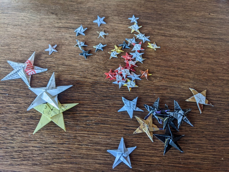 Puede incluir: Estrellas de origami de varios tama&ntilde;os y colores, incluyendo blanco, amarillo, rojo y negro. Las estrellas est&aacute;n dispuestas sobre una superficie de madera, mostrando los intrincados pliegues y patrones del papel.