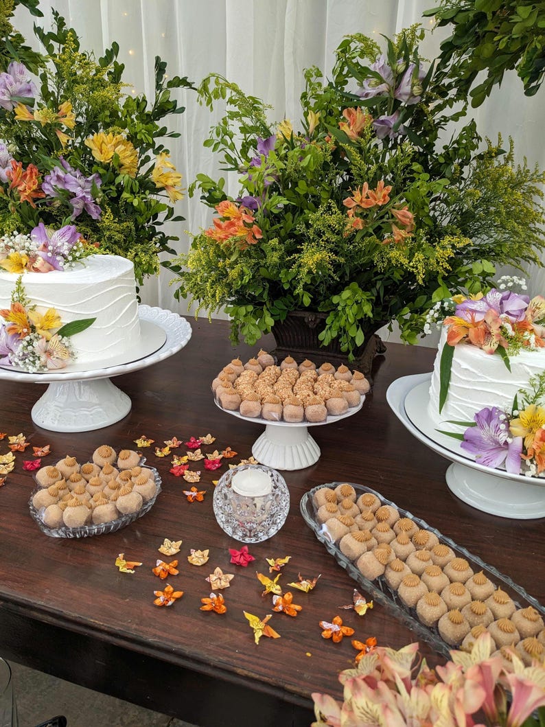 May include: A table setting with two white cakes decorated with flowers, surrounded by small brown and white candies, and a clear glass candle holder. The table is decorated with origami paper butterflies in shades of orange, yellow, and red.