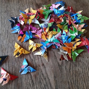 May include: A collection of colorful origami butterflies made from recycled materials. The butterflies are arranged in a pile on a wooden surface.