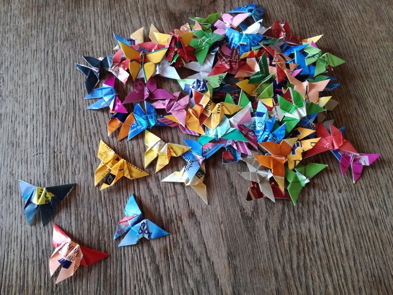 May include: A collection of colorful origami butterflies made from recycled materials. The butterflies are arranged in a pile on a wooden surface.