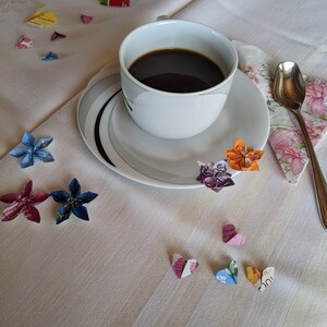 May include: A white coffee cup with a saucer and a spoon on a white tablecloth with colorful origami paper flowers and hearts.