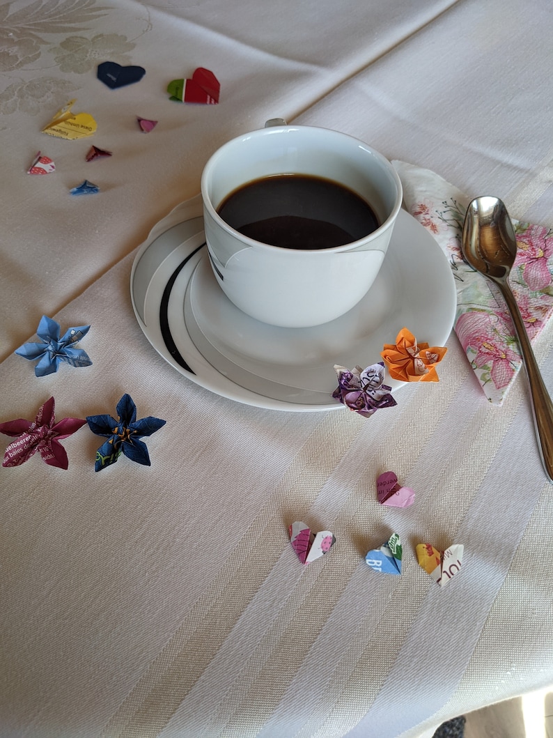 May include: A white coffee cup with a saucer and a spoon on a white tablecloth with colorful origami paper flowers and hearts.