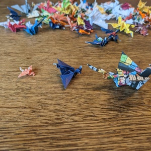 Może przedstawiać: Kolekcja kolorowych żurawi origami z papieru, niekt&oacute;re złożone w r&oacute;żne kształty, na drewnianej powierzchni. Na pierwszym planie znajdują się trzy żurawie, jeden pomarańczowy, jeden niebieski i jeden z kolorowym wzorem komiksowym.