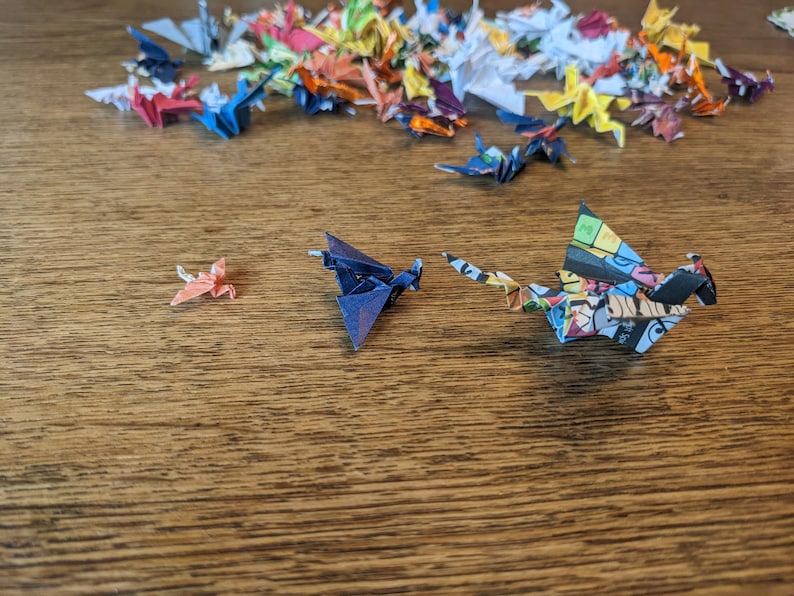 Może przedstawiać: Kolekcja kolorowych żurawi origami z papieru, niekt&oacute;re złożone w r&oacute;żne kształty, na drewnianej powierzchni. Na pierwszym planie znajdują się trzy żurawie, jeden pomarańczowy, jeden niebieski i jeden z kolorowym wzorem komiksowym.