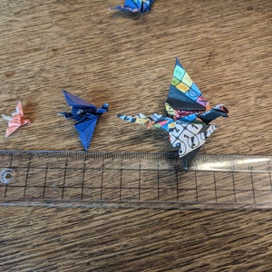 Może przedstawiać: Trzy origami z papieru w kształcie żurawi, jeden r&oacute;żowy, jeden niebieski i jeden wielokolorowy, są ułożone na drewnianej powierzchni z linijką jako skalą. Żuraw wielokolorowy jest największy, po nim następuje żuraw niebieski, a najmniejszy jest żuraw r&oacute;żowy.
