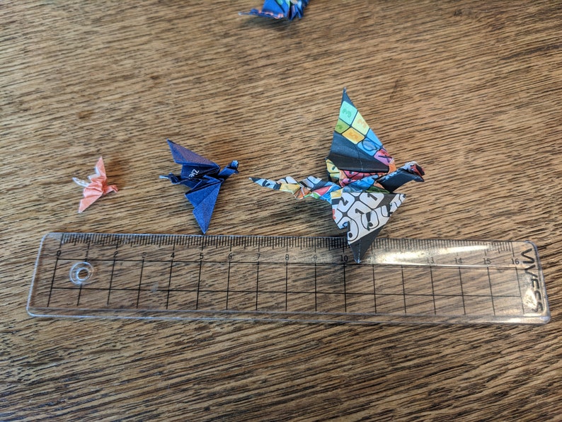 Może przedstawiać: Trzy origami z papieru w kształcie żurawi, jeden r&oacute;żowy, jeden niebieski i jeden wielokolorowy, są ułożone na drewnianej powierzchni z linijką jako skalą. Żuraw wielokolorowy jest największy, po nim następuje żuraw niebieski, a najmniejszy jest żuraw r&oacute;żowy.
