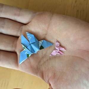 May include: Two origami butterflies, one blue and one pink, are folded from paper and displayed on a person's hand.
