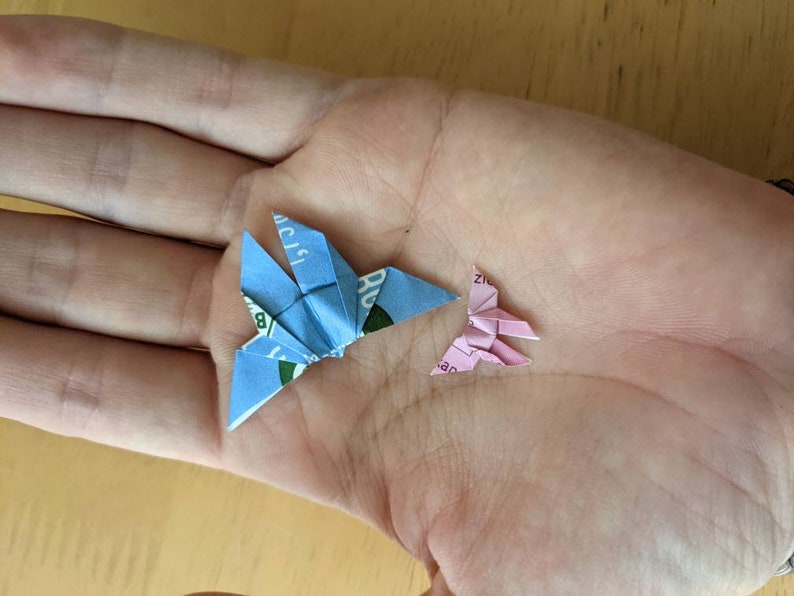 May include: Two origami butterflies, one blue and one pink, are folded from paper and displayed on a person's hand.