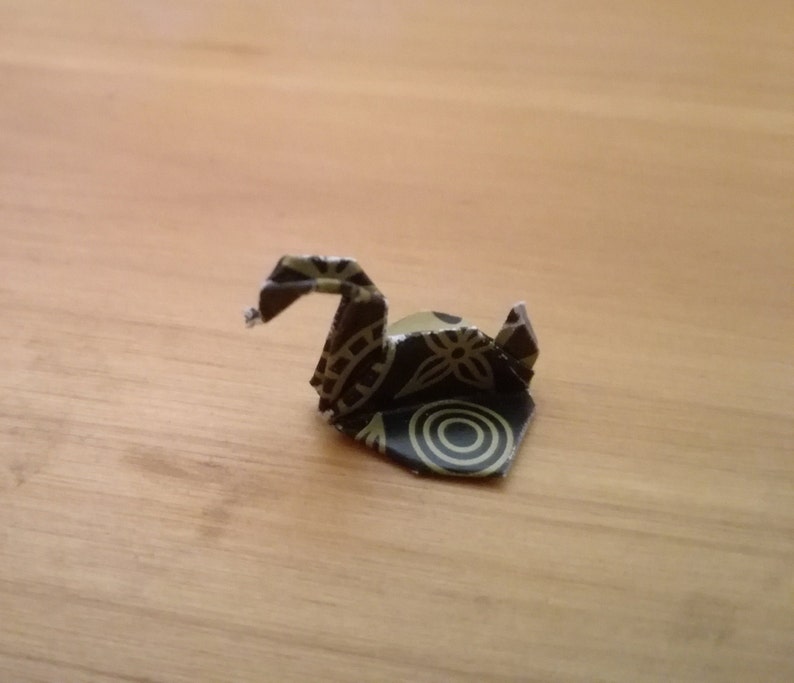 May include: A small origami swan made from paper with a black and gold floral pattern.
