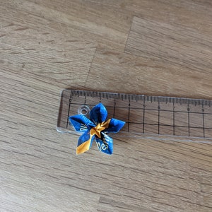 May include: A blue and yellow origami flower with a star in the center. The flower is about 2 inches wide and is sitting on a ruler.
