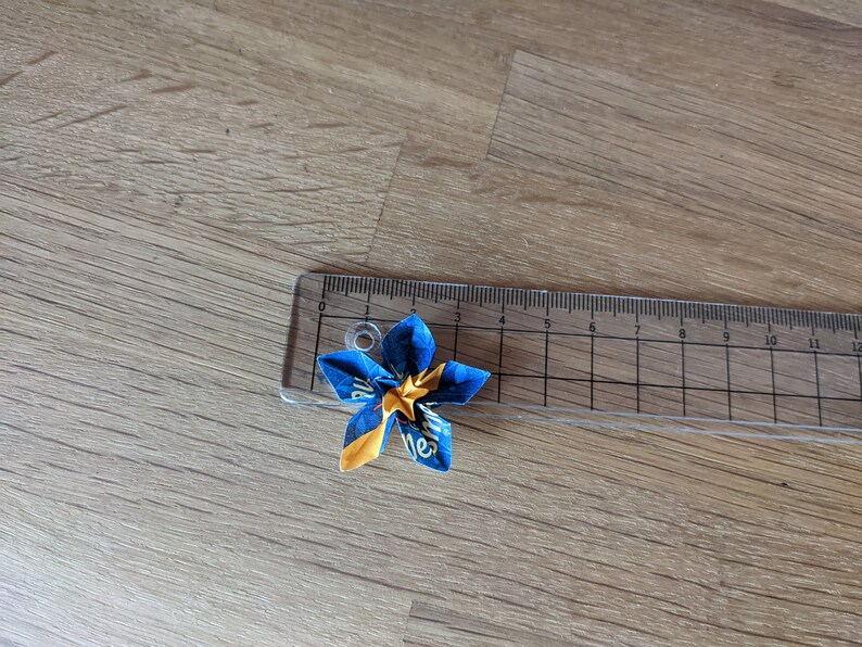 May include: A blue and yellow origami flower with a star in the center. The flower is about 2 inches wide and is sitting on a ruler.
