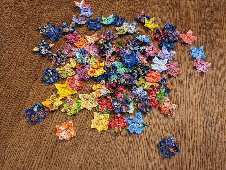 May include: A collection of colorful origami flowers made from paper. The flowers are in various shades of blue, red, yellow, green, and purple. They are arranged in a cluster on a wooden surface.