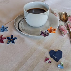 May include: A white coffee cup with a black saucer and a cup of coffee. There are several origami flowers and hearts on a white tablecloth. The text on one of the hearts reads "something special".