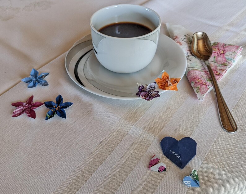 May include: A white coffee cup with a black saucer and a cup of coffee. There are several origami flowers and hearts on a white tablecloth. The text on one of the hearts reads "something special".