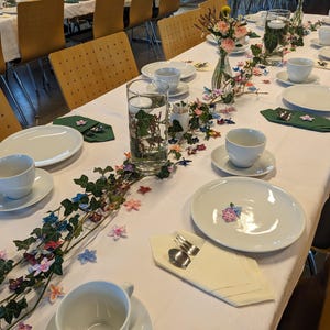 May include: A white tablecloth with a green ivy garland and colorful paper flowers. There are white plates, cups, and saucers set for a dinner party.