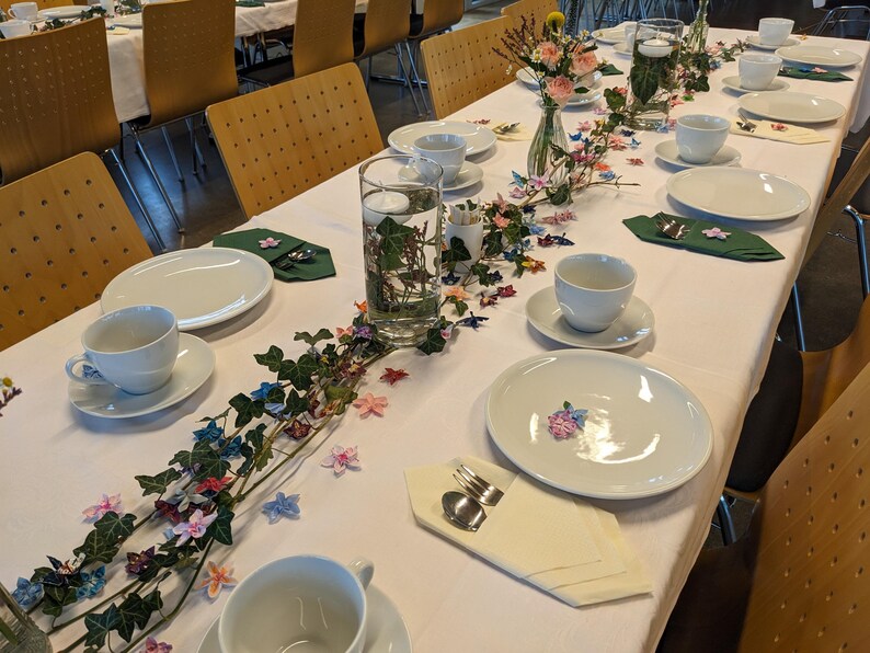 May include: A white tablecloth with a green ivy garland and colorful paper flowers. There are white plates, cups, and saucers set for a dinner party.