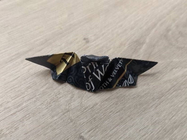 Puede incluir: Una figura de origami plegada con papel estampado en negro y dorado. El papel tiene el texto "Prince of Wales" y "Smooth & Velvety". El origami est&aacute; sobre una superficie de vetas de madera clara.