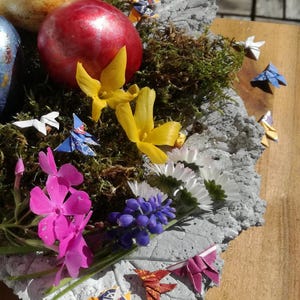 May include: A concrete nest with moss, decorated with origami birds, flowers, and painted Easter eggs. The nest is on a wooden surface.